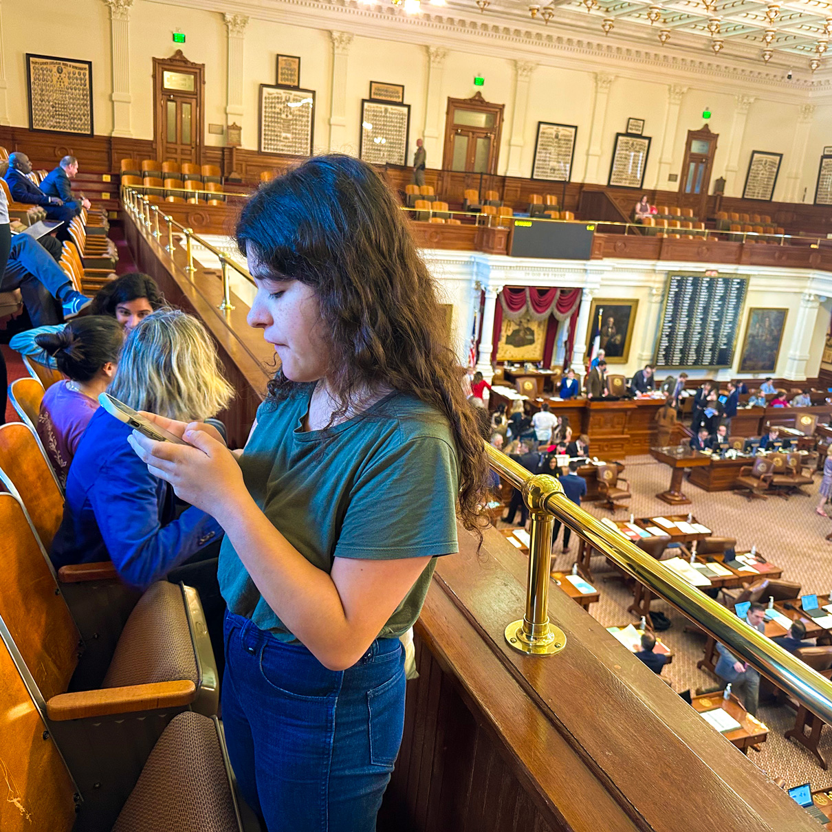 University student stands in the gallery of the Texas House of Representatives, with the brass railing of the gallery to her back as she checks her cellphone for networking messages.